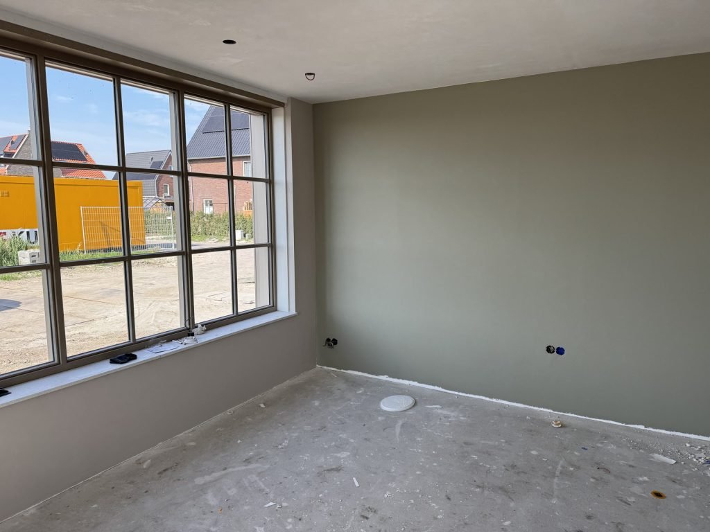 Nieuwbouwkamer met een egaal afgewerkte grijsgroene wand en een groot raam met zicht op woningen in aanbouw