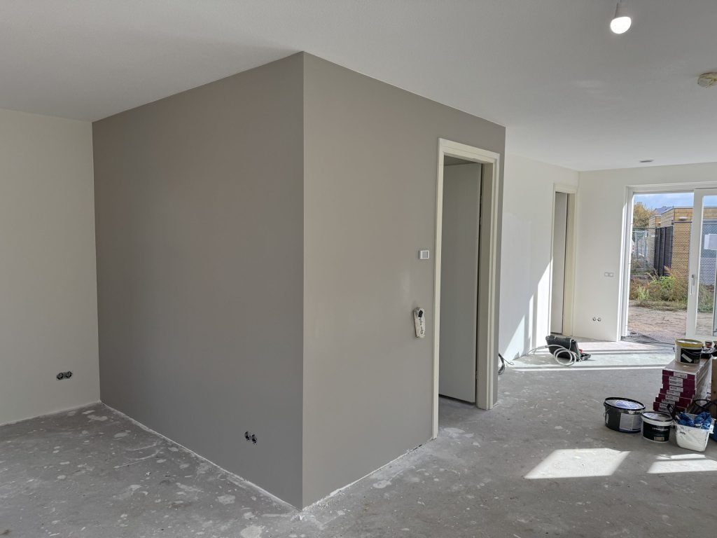 Open woonkamer in een nieuwbouwhuis met een net afgewerkte grijze wand en verfemmers op de dekvloer